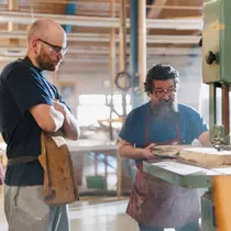 Two men working together in a woodshop, one holding a piece of wood while the other operates a machine.